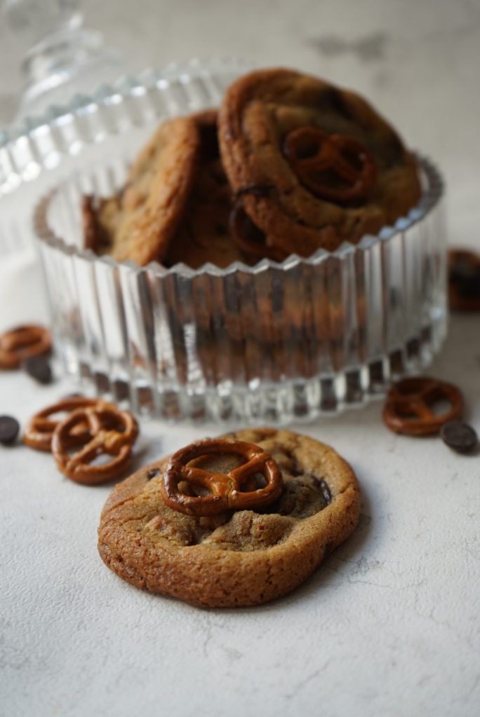 Een close-up van een koekje met chocolade en een pretzel erop, naast een glazen koekjespot en kleine chocoladeschijfjes.