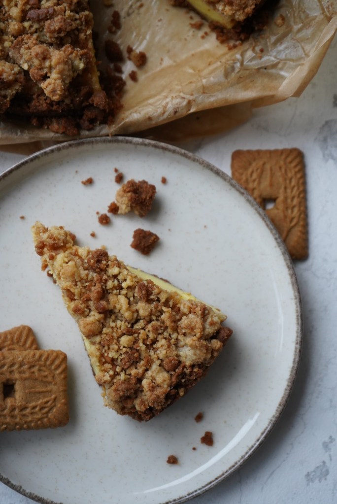 Een stuk cheesecake met een kruimellaag op een wit bord, naast enkele speculaasjes.